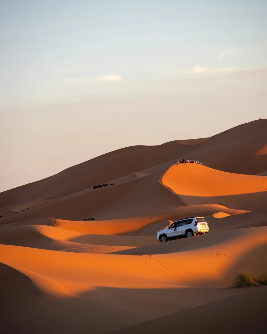 A white 4x4 driving across the sunlit dunes of Erg Chebbi